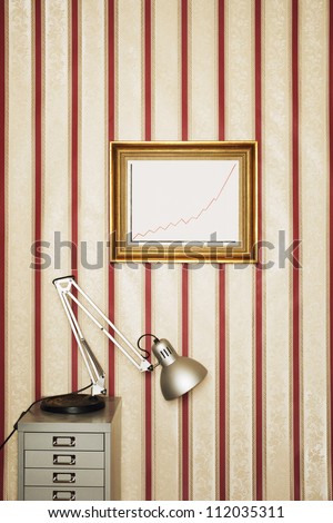 Lamp on file cabinet and framed graph on patterned wallpaper in office