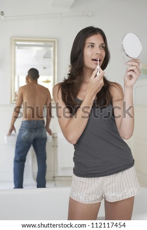 Beautiful woman applying lipstick with man in the background