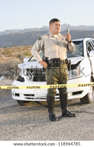 Police officer using radio in front of damaged car in desert
