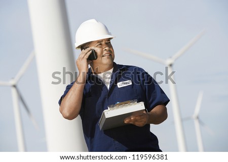 Male engineer on call holding clipboard and looking away at wind farms