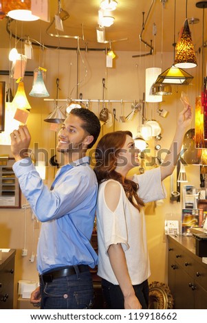 Happy young couple standing back to back while looking at price tag in lights store