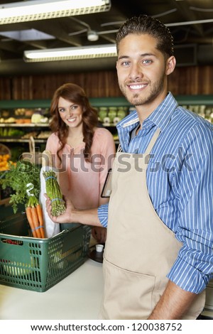 Happy young sales clerk holding vegetables with woman in background