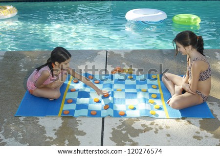 Two girls playing large draughts board beside swimming pool