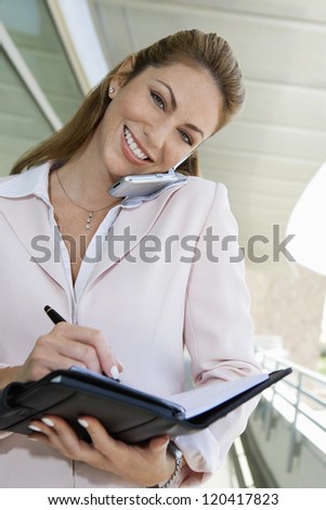 Portrait of a happy Caucasian businesswoman writing notes while communicating on mobile phone