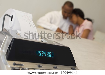 Closeup of expense receipt machine with tensed African American couple in the background