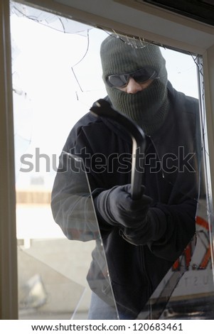 Thief in balaclava breaking glass of window to enter the house