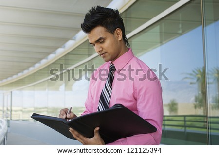 An Indian businessman writing notes in folder while standing at office balcony