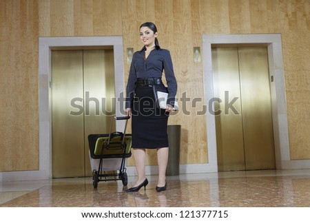 Full length of a happy business woman pulling trolley against elevators