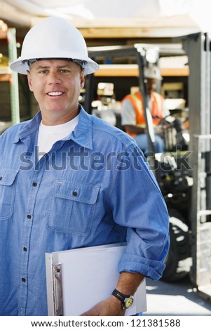 Portrait of a male industrial worker holding clipboard with coworker in the background