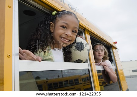Portrait of little girl looking out from window of school bus with classmate in background