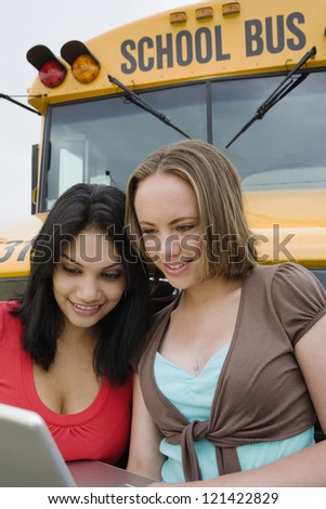 Happy young female friends using laptop against school bus