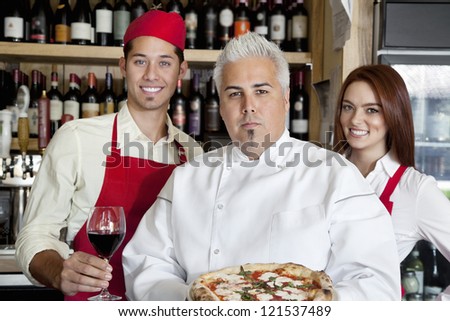 Portrait of a confident chef holding pizza with wait staff in background