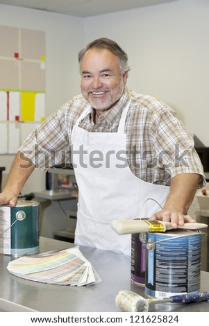 Portrait of a cheerful middle-aged store clerk with paint cans and brush in hardware shop