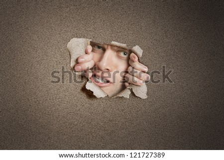 Portrait of a aggressive young man peeking from ripped white paper hole