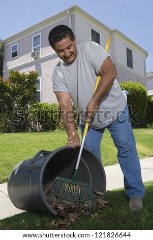 Hispanic man sweeping leaves into a trash with house in background
