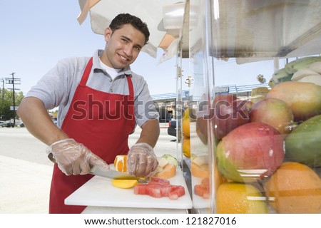 Portrait of young hispanic male street vendor chopping fruits on board