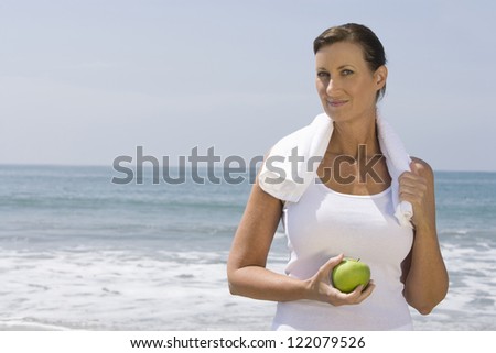 Portrait of sporty woman holding green apple with towel around shoulders on beach