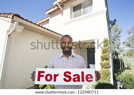 Portrait of an African American mature estate agent holding \'For Sale\' sign against house