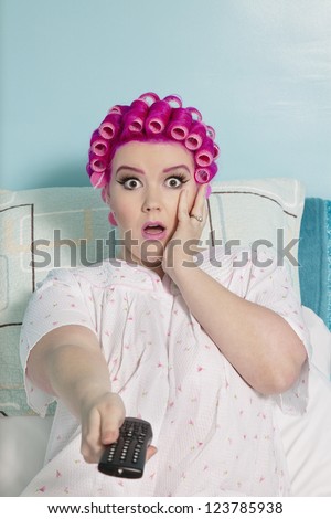 Portrait of shocked woman holding remote with hair curlers sitting on bed