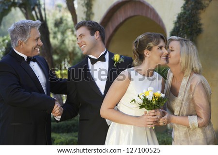 Mother in law kissing bride with happy father and son shaking hands in the background