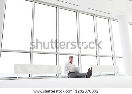 Full length of young businessman using laptop while sitting amidst radiators against window at office