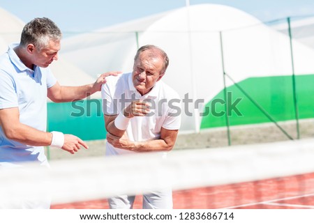 Mature man helping senior friend with elbow injury while playing tennis on sunny day