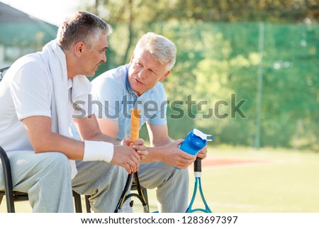 Mature male friends talking while sitting with tennis rackets and water bottle on chair at court