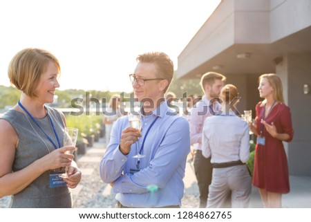 Businessman have an intellectual conversation with his secretary on the balcony at their corporate business retreat they are both sharing a glass of wine and their colleagues are in the background