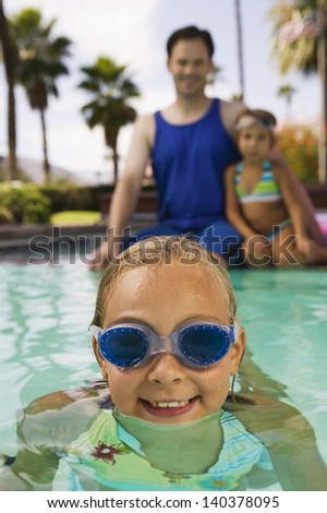 Girl in swimming pool with father and sister sitting at pool edge in background