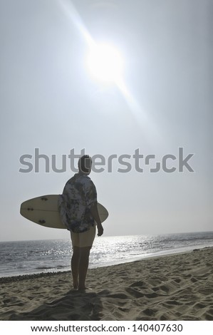 Rear view of a man in Hawaiian shirt with surfboard standing on beach