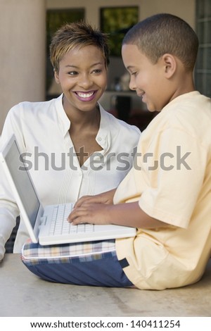 Boy sitting on counter and using laptop with happy mother in the living room