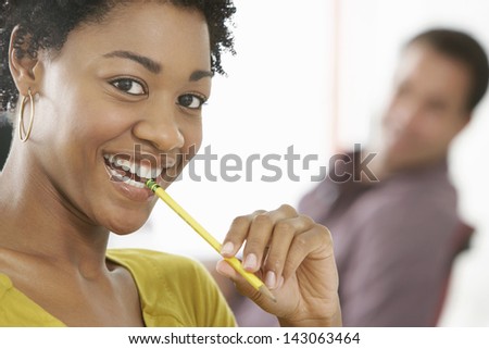 Portrait of smiling young businesswoman chewing pencil with colleague in background
