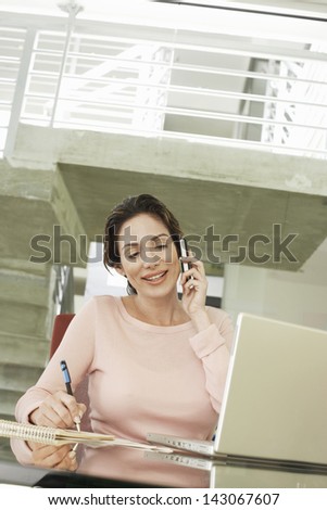 Happy young businesswoman using mobile phone while writing on notepad in office