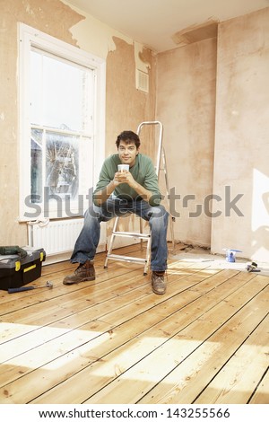 Full length portrait of man sitting on step ladder while holding coffee mug in unrenovated room