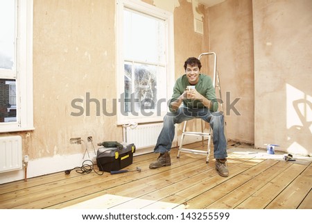 Full length portrait of happy man sitting on step ladder while holding coffee mug in unrenovated room