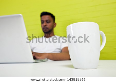Indian man working at his desktop with mug in focus in foreground