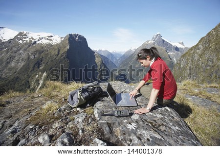 Side view of a young woman using laptop on boulder against mountains