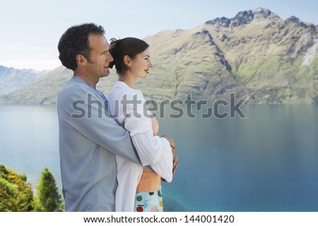 Side view of a man embracing woman from behind while looking at the mountain lake