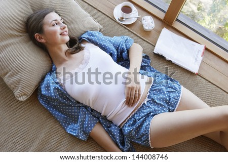 Young woman in pajamas lying on couch with coffee and magazine on window sill