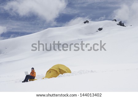 Side view of a male hiker using laptop by tent on snowy mountain slope