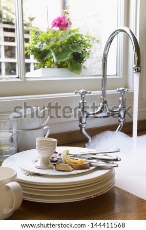 Closeup Of A Stack Of Dirty Dishes And Silverware By Sink With Running Water Below Window With Flower