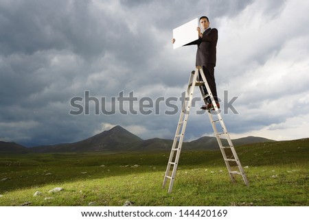 Full length of businessman on ladder holding blank sign in mountain field