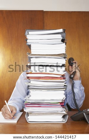 Obscured office worker using telephone while writing document behind a stack of folders at desk