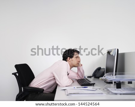 Side View Of A Bored Male Office Worker Looking At Notes On Computer Monitor At Desk