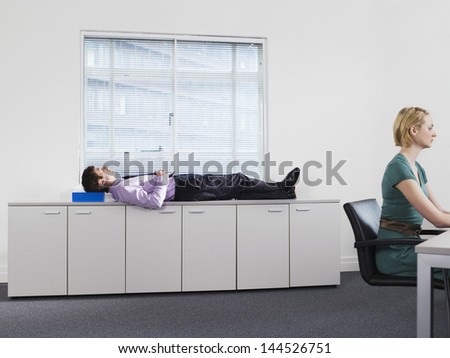 Businessman sleeping on office cabinets while woman working in foreground
