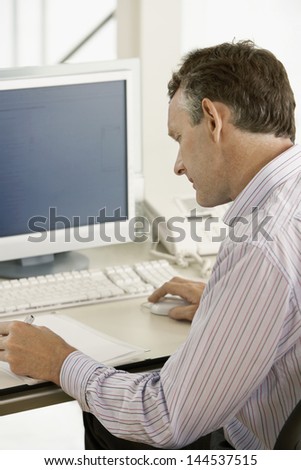 Middle aged male businessman using computer while noting on paper at desk