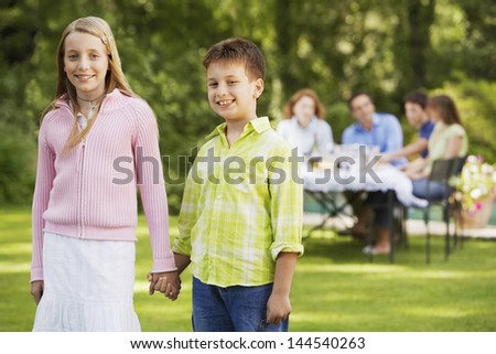 Portrait of happy brother and sister holding hands with family having lunch in background