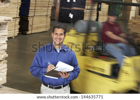 Portrait of a smiling confident man with clipboard in front of forklift in warehouse