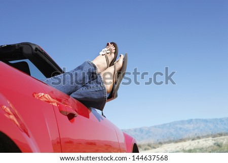 Woman resting in red convertible with legs sticking out against clear blue sky
