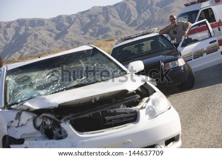 Police officer using radio in front of a damaged car on desert road